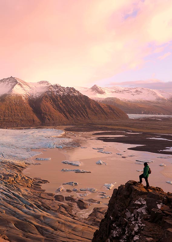 A hiker stands at the edge of a cliff overlooking Icelandic mountains in sunset.